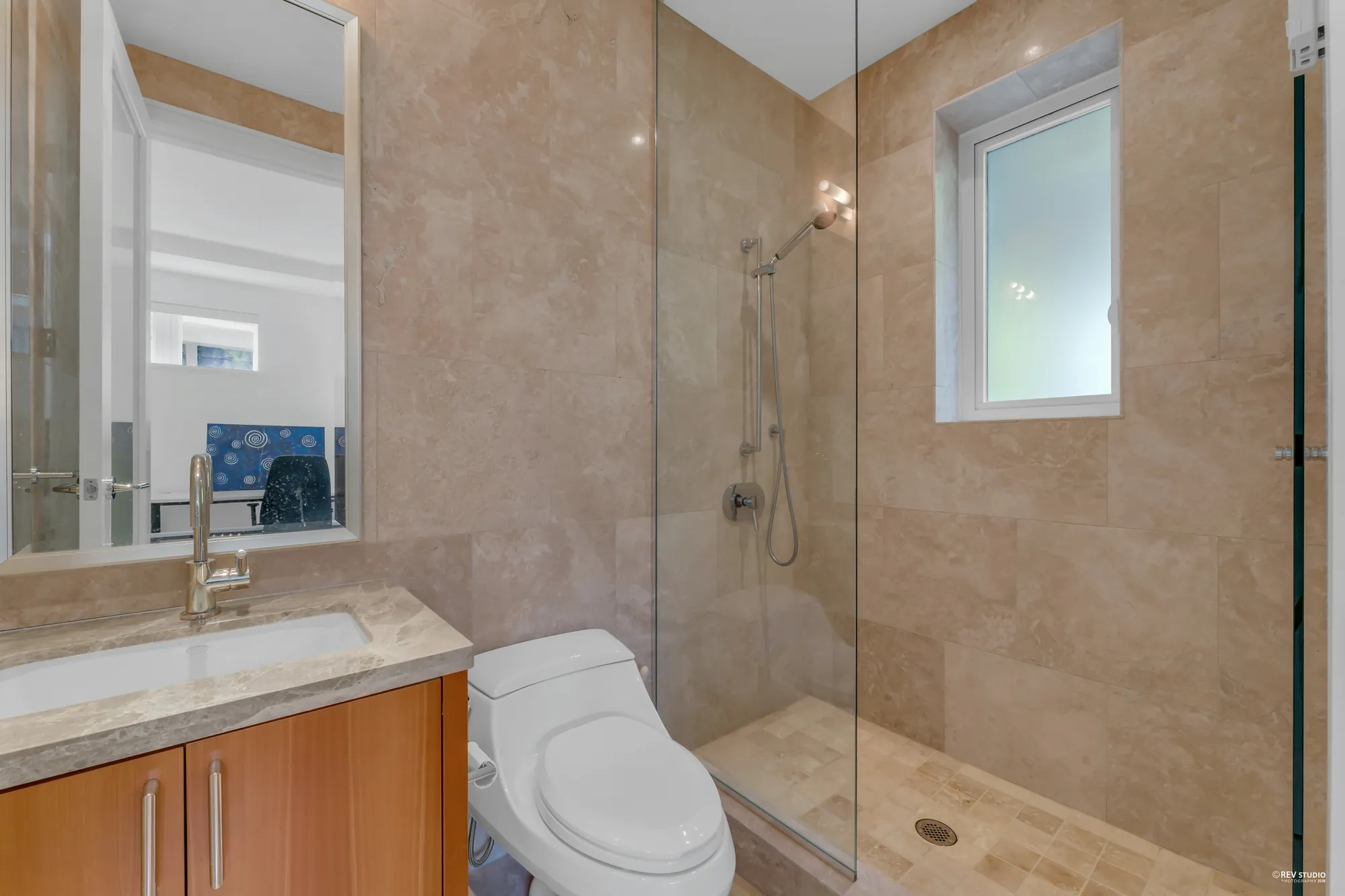 The second bathroom: a wood vanity with a stone counter and a brass-coloured tap, a glass shower tiled in beige stone, a window