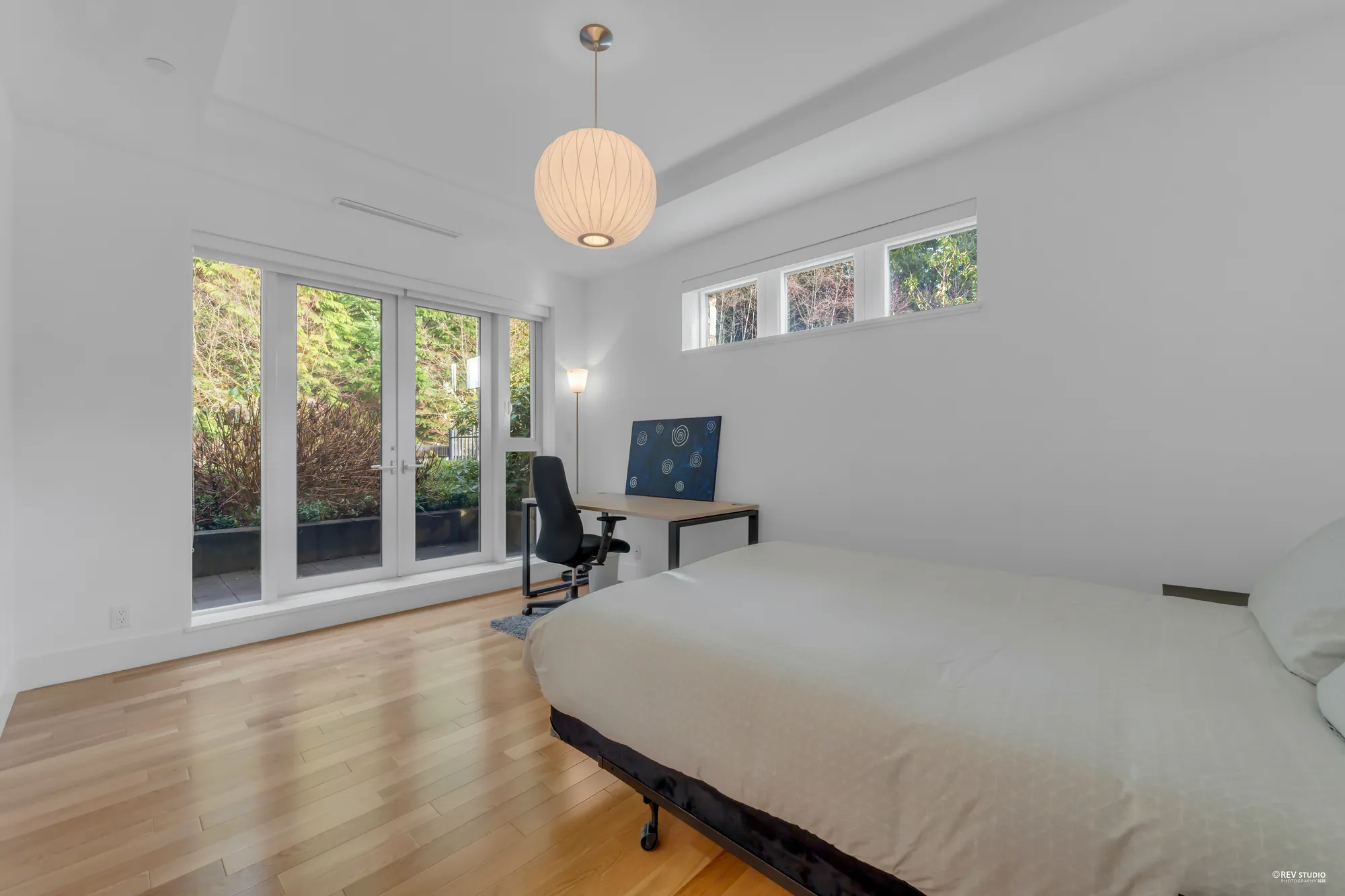 The second bedroom: a bed, a small desk and chair, a round paper pendant, French doors out to the planting and a row of high windows