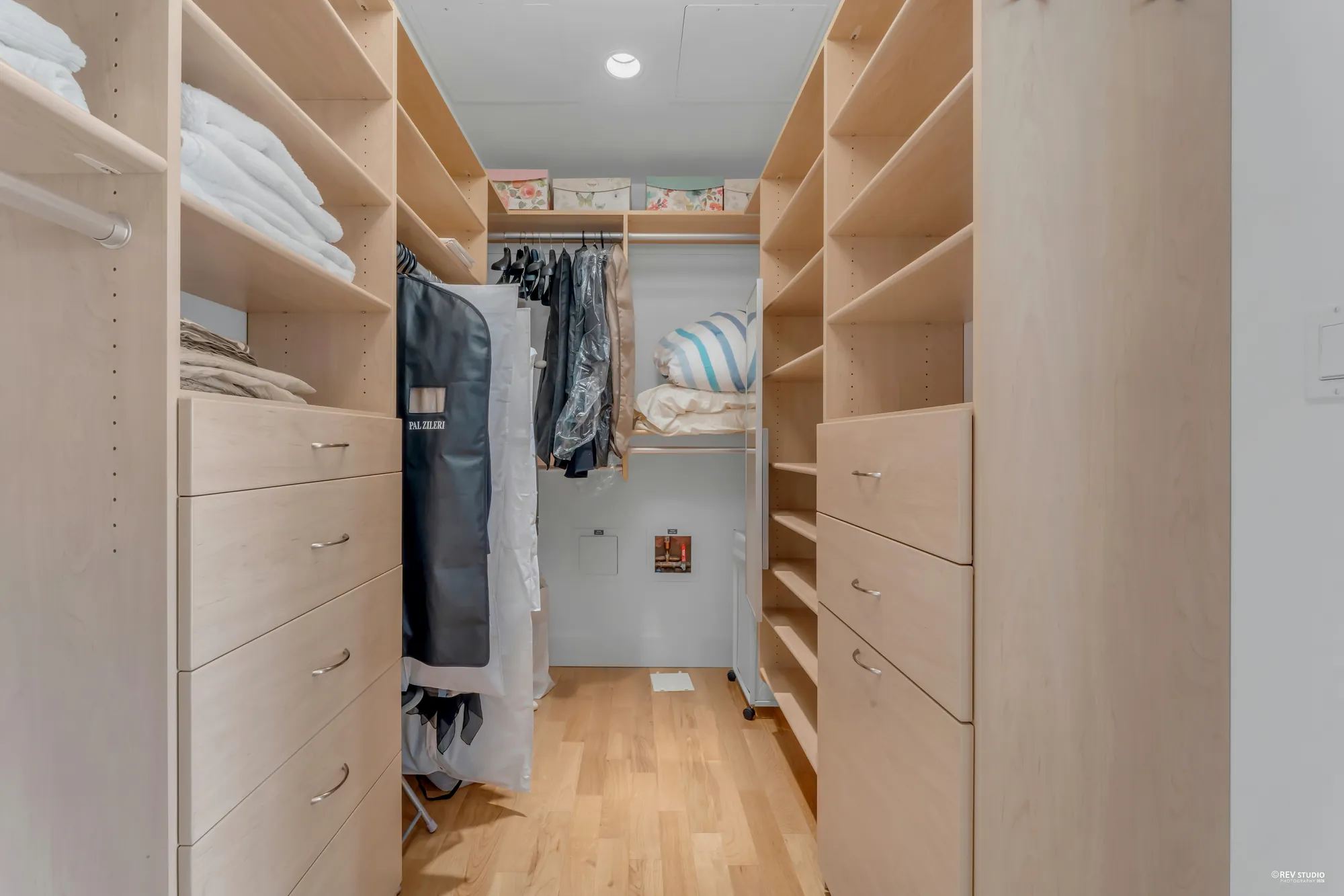 The walk-in closet: pale wood drawers and shelving on both sides and hanging rails at the end