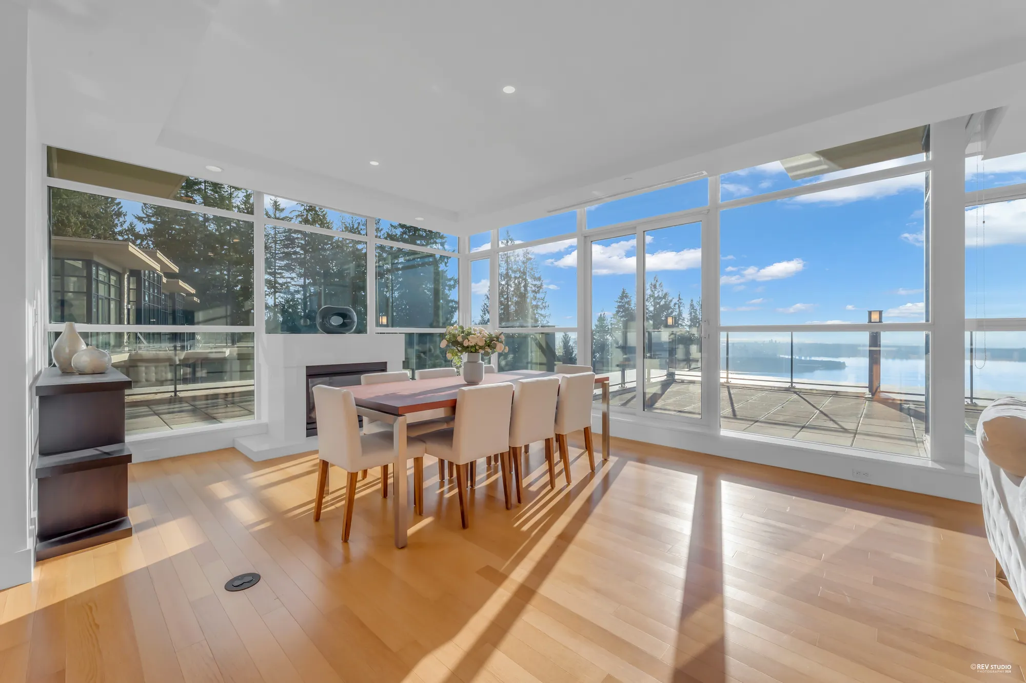 The dining area: a wood table with white chairs by the windows, a white fireplace at the left, the terrace and the water beyond