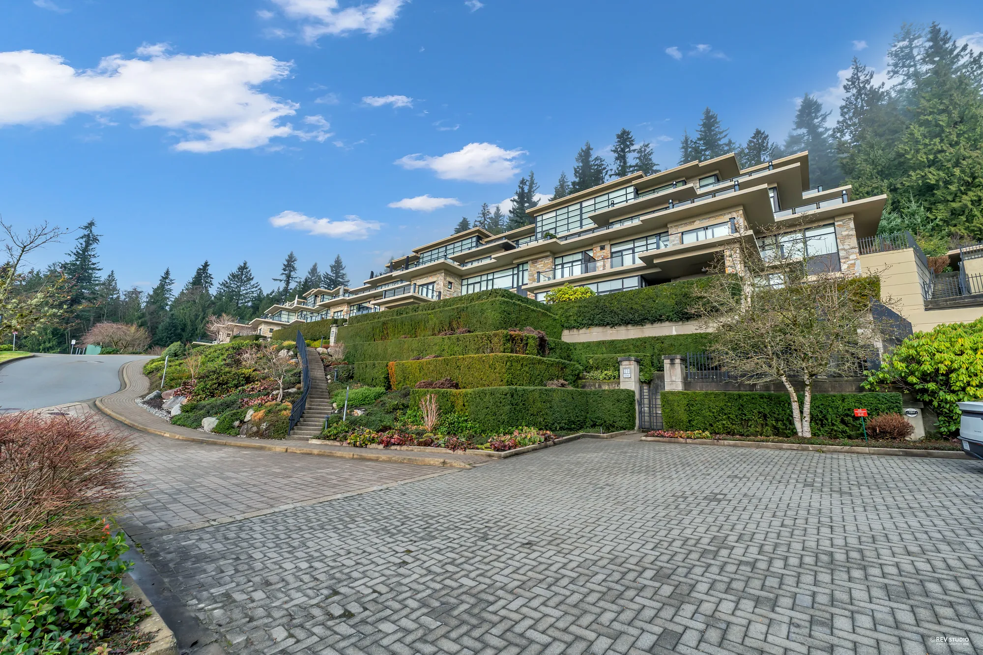The terraced buildings from the paved drive, a planted slope of shrubs and boulders below them