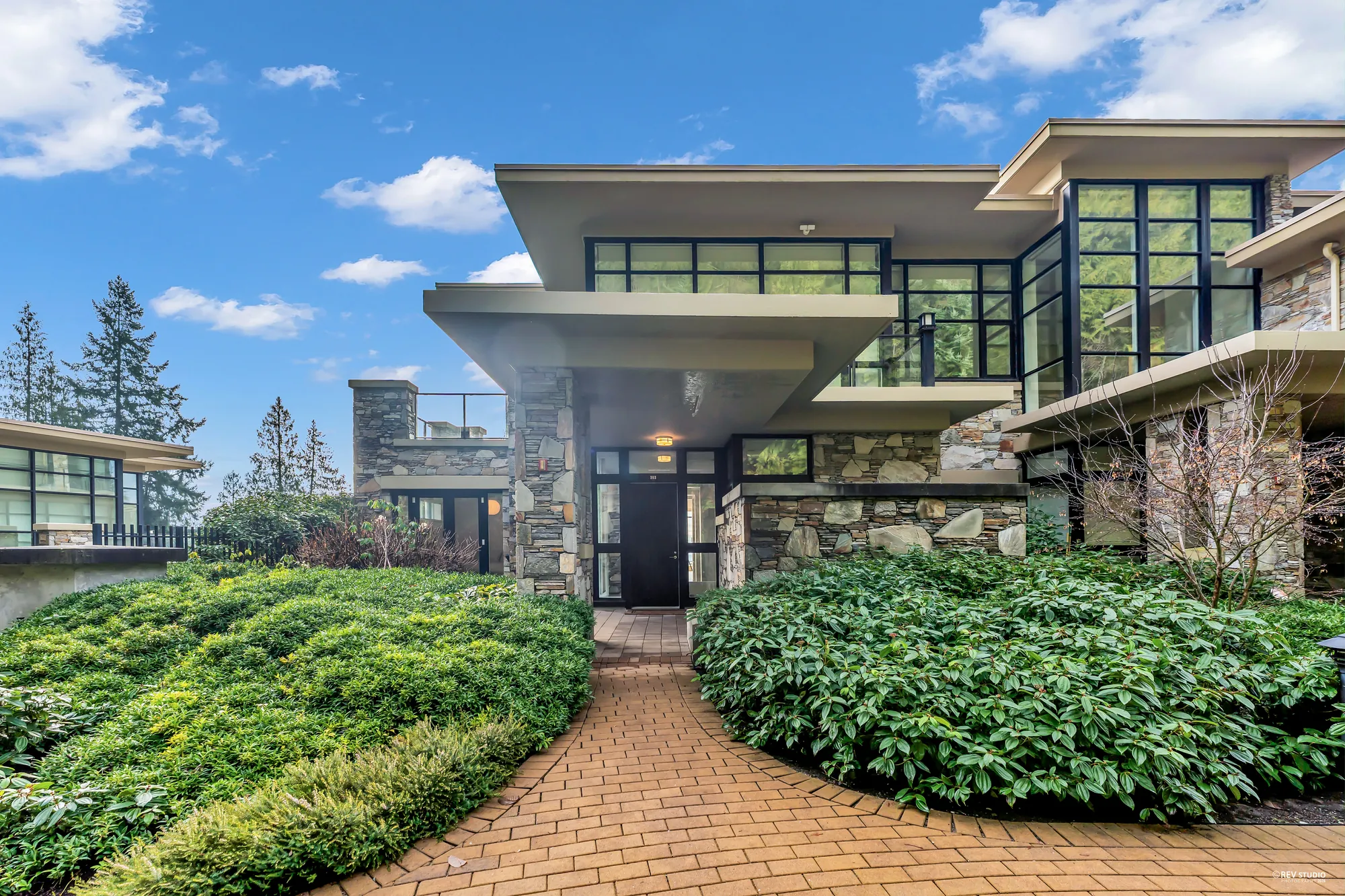 A straight brick path between rounded shrubs to a glass door under a deep overhang, stone piers on either side