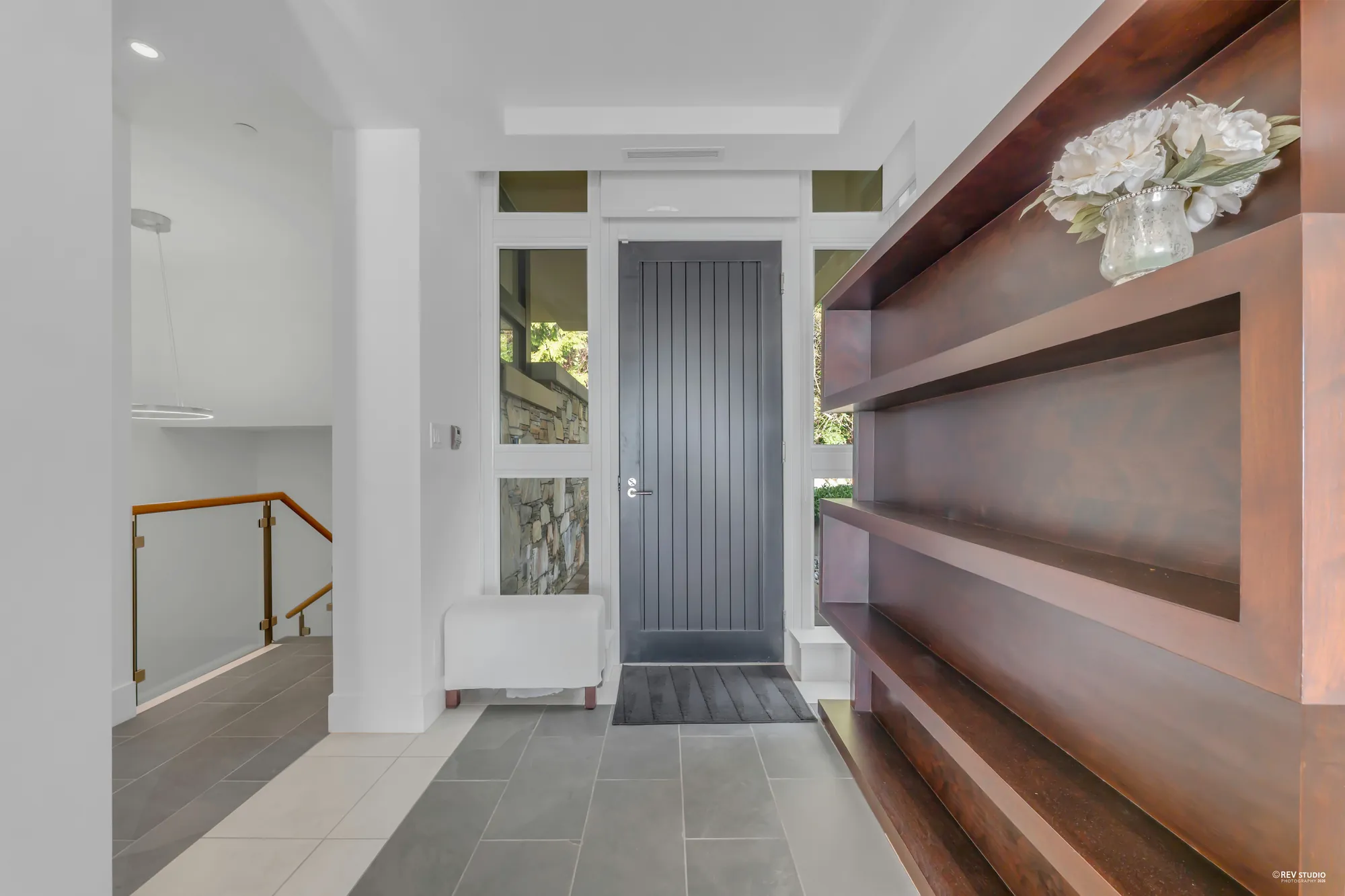 The foyer: a tall dark front door with glass sidelights, a grey tile floor, a stair with a glass rail going down at the left and a wood shelf unit at the right