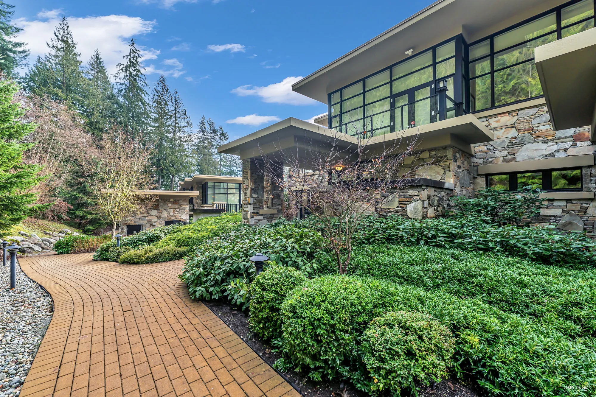 The brick path winding through clipped shrubs and garden beds beside the stone building, evergreens beyond