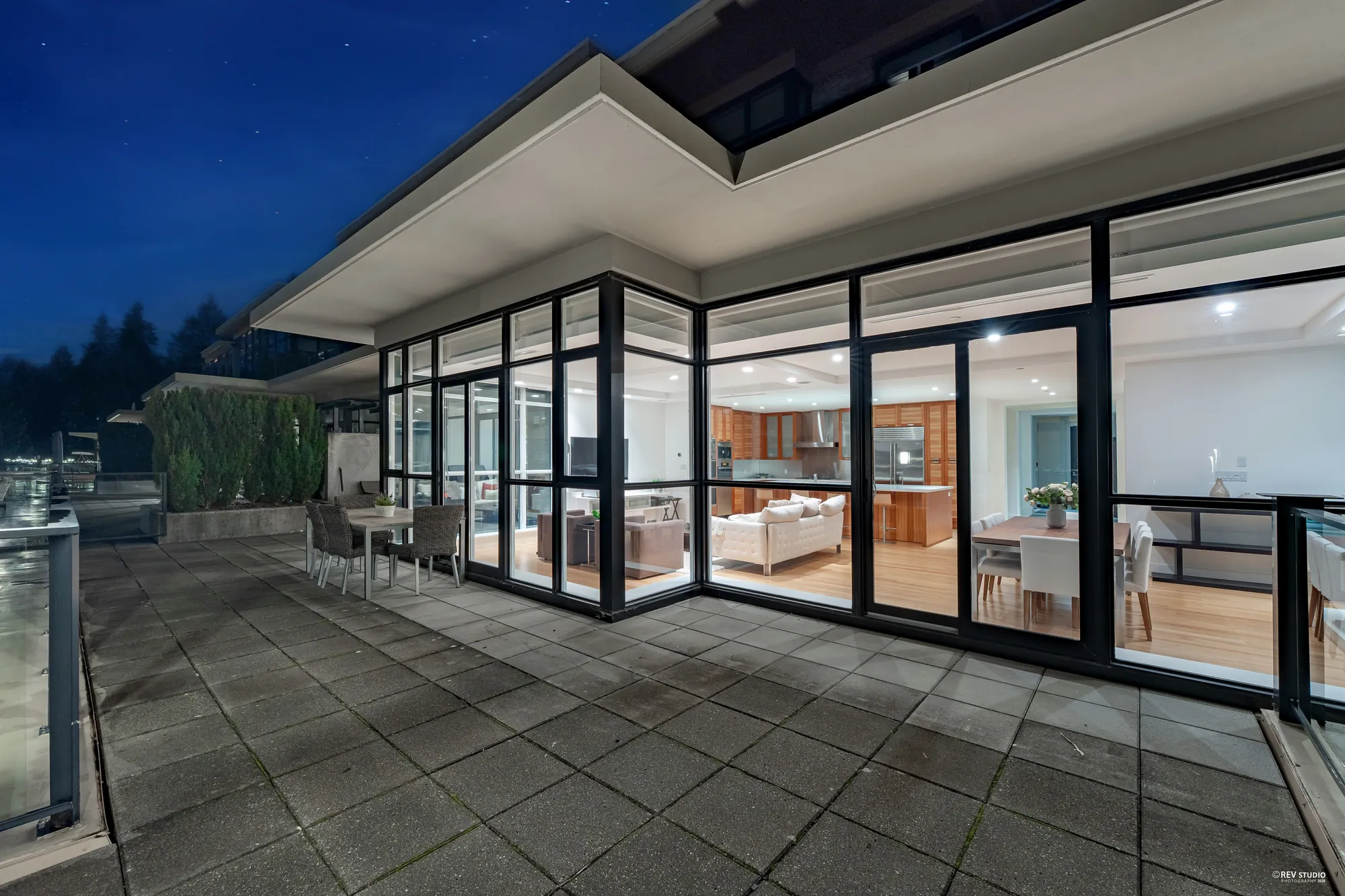 The living room and the kitchen lit behind a wall of glass, seen from the terrace at dusk