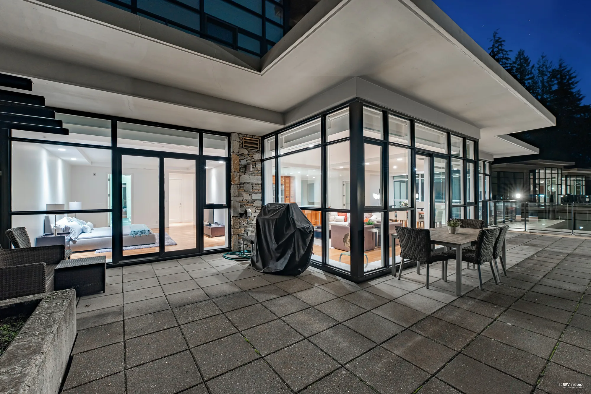 The home from the terrace at dusk: rooms lit behind a corner of glass, stone at the corner, a covered barbecue and patio chairs