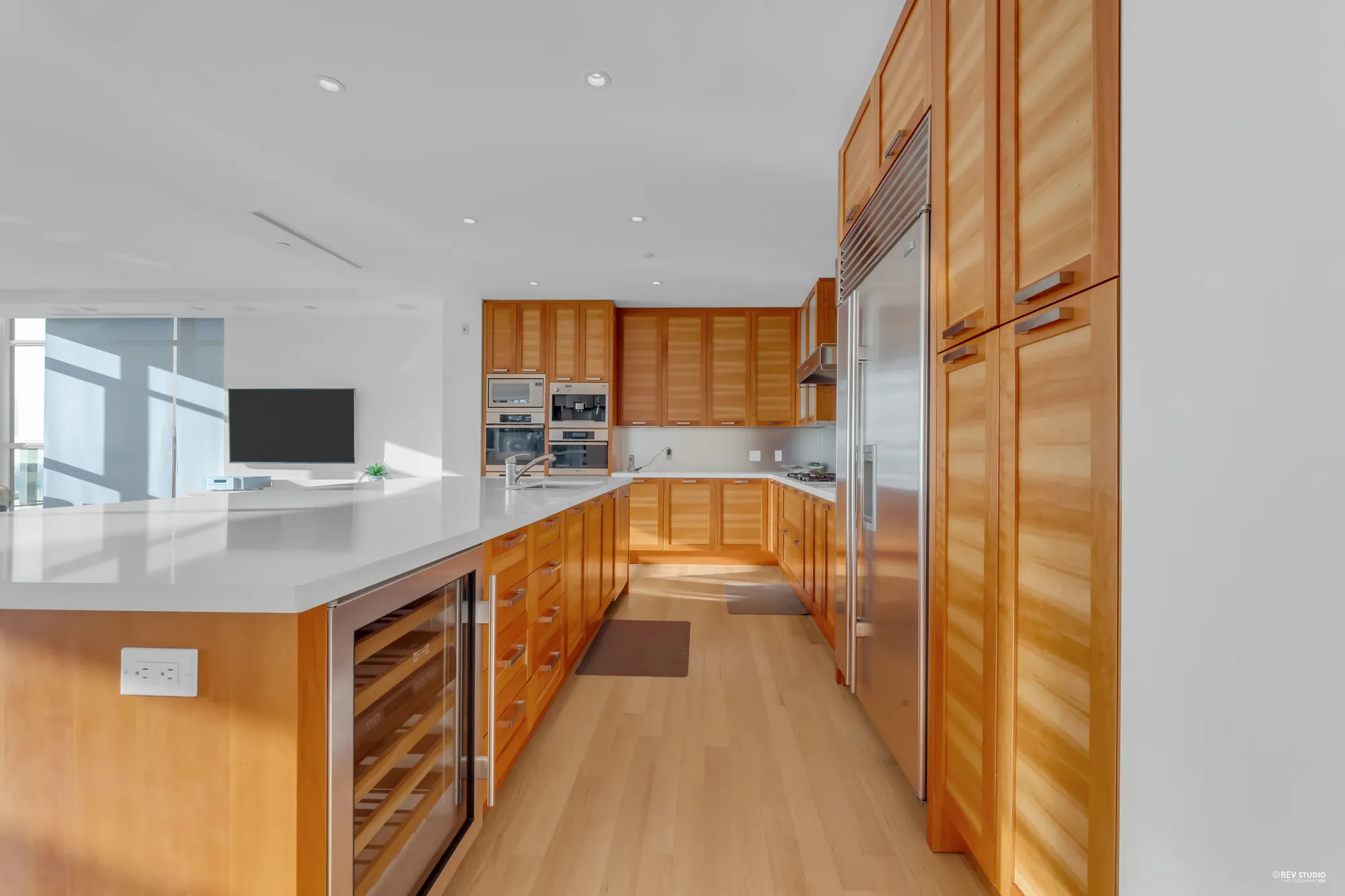 Along the kitchen: tall wood panels around the built-in fridge, wall ovens, wood cabinets, a wine fridge at the end of the island and the windows at the far end