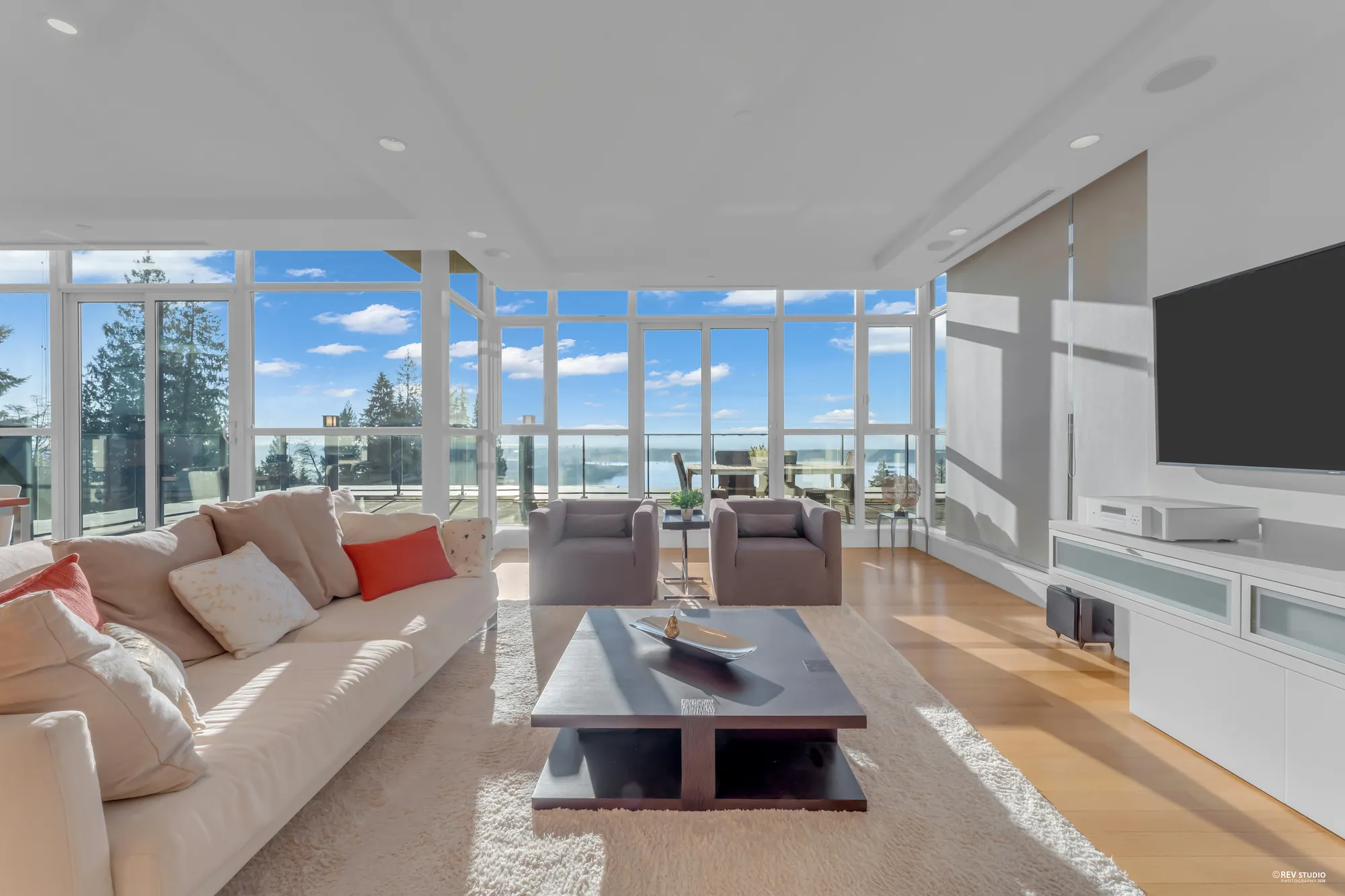The living room by day: a cream sofa with orange cushions, two grey armchairs and a dark coffee table on a pale rug, windows from floor to ceiling on two sides with the water beyond, light wood floors