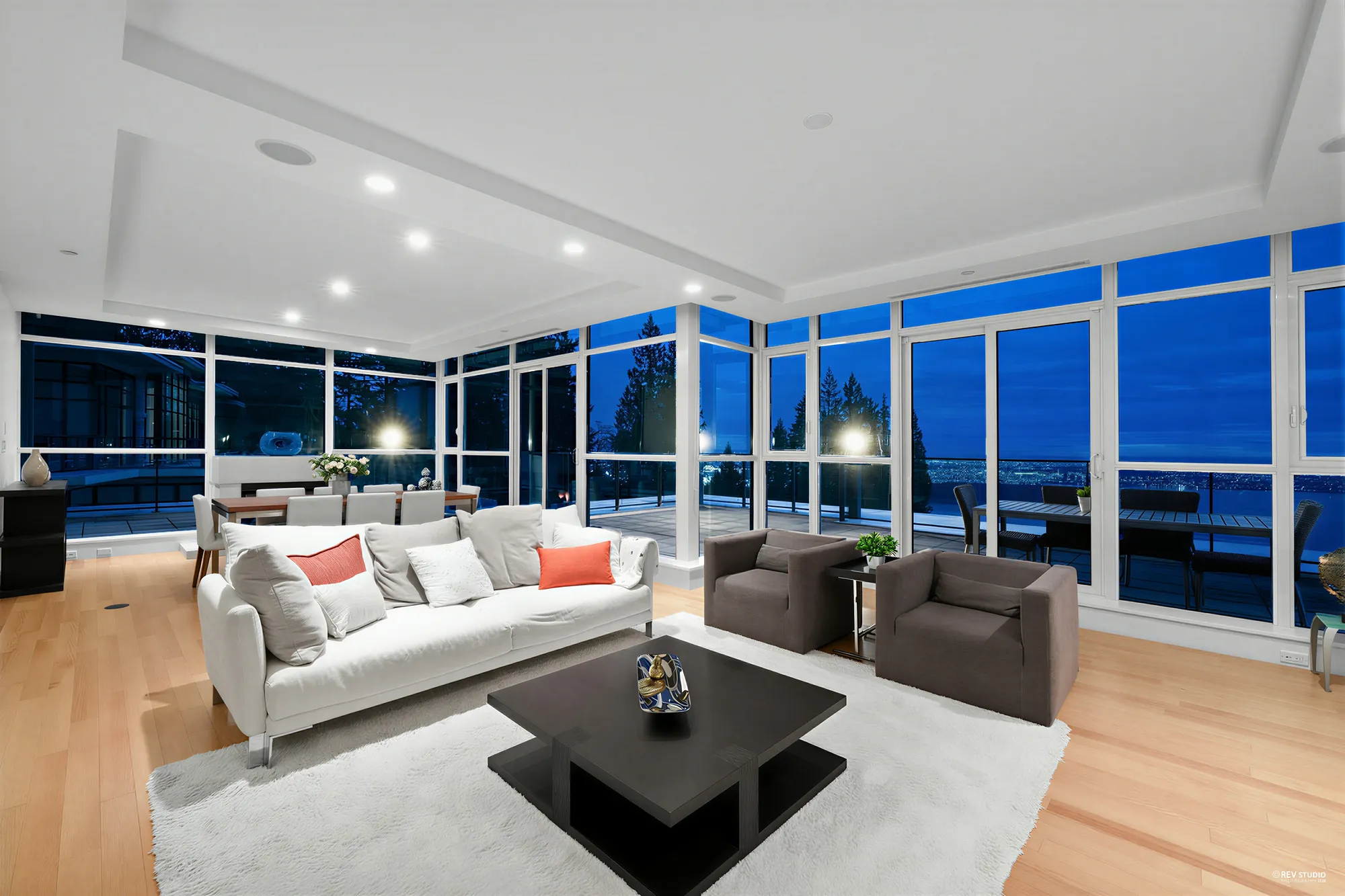 The living room from the same corner at dusk, the lamps and ceiling lights on, the terrace outside the windows