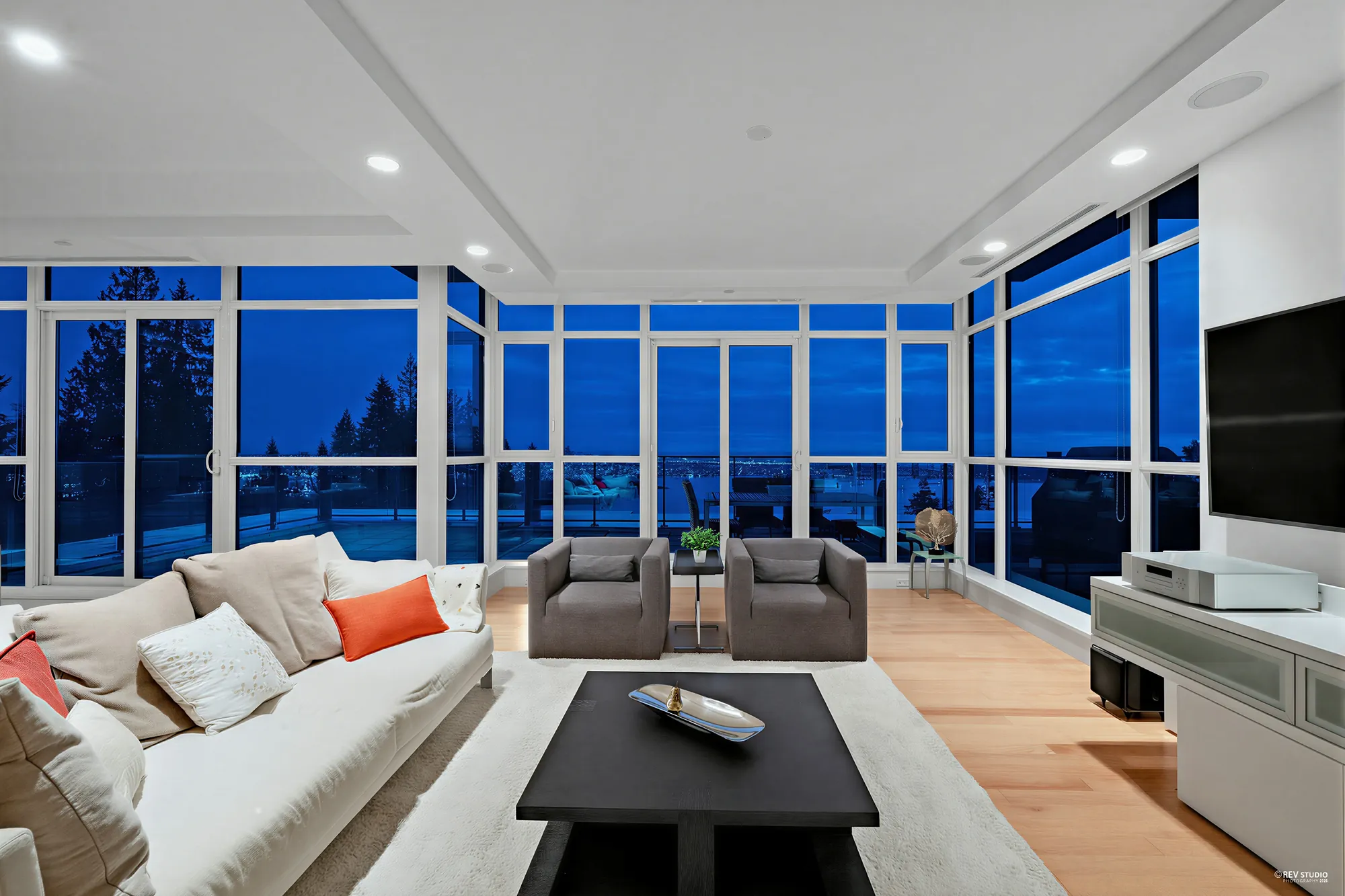 The same corner of the living room at dusk, the windows deep blue and lights along the far shore of the water