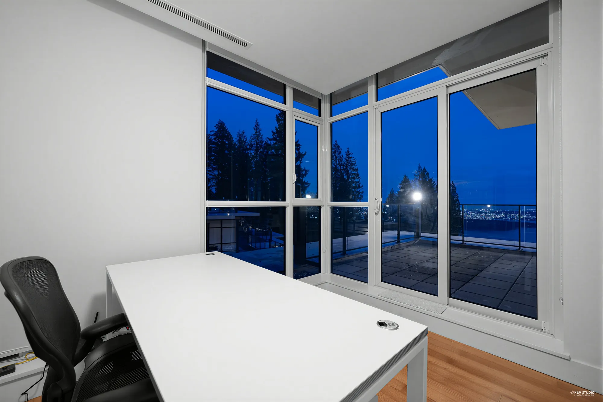 The office at dusk: a white desk and a black office chair in a corner of windows from floor to ceiling, the balcony and trees outside
