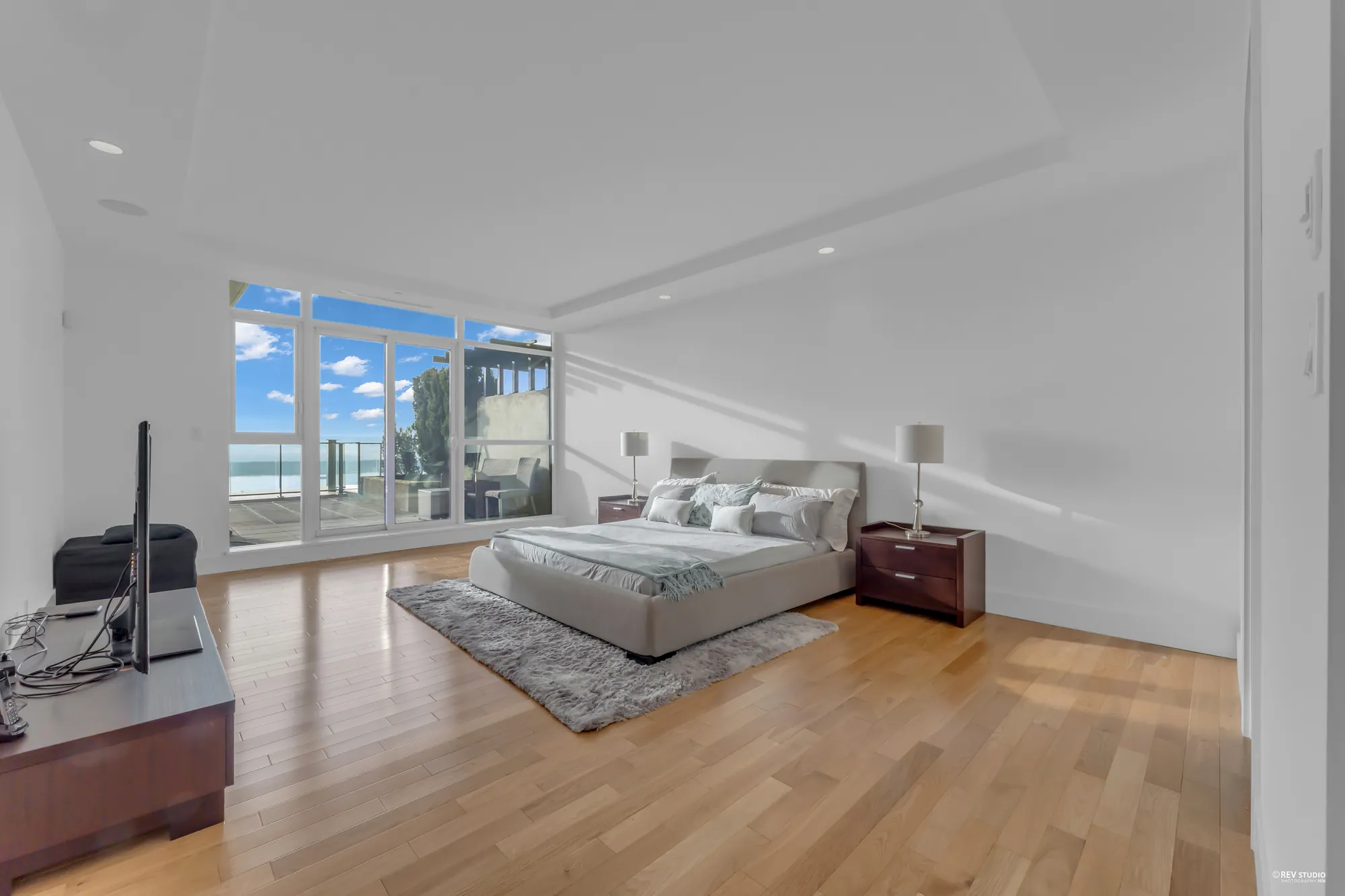 The primary bedroom: a grey upholstered bed on a rug, two night tables with lamps, windows along the far wall with the water beyond, light wood floors