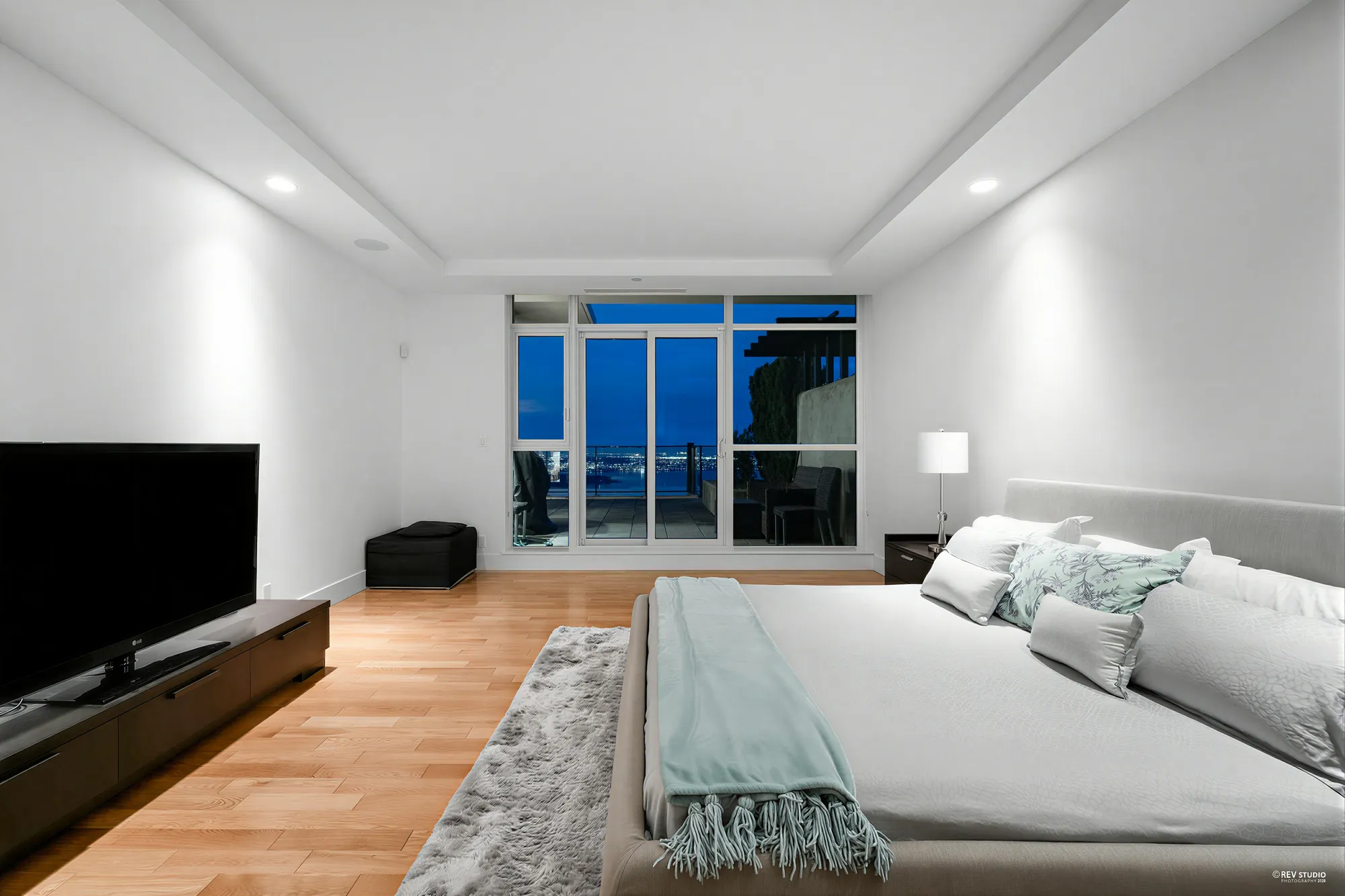 The primary bedroom at dusk from beside the bed, a television on a low cabinet, the windows and the view beyond