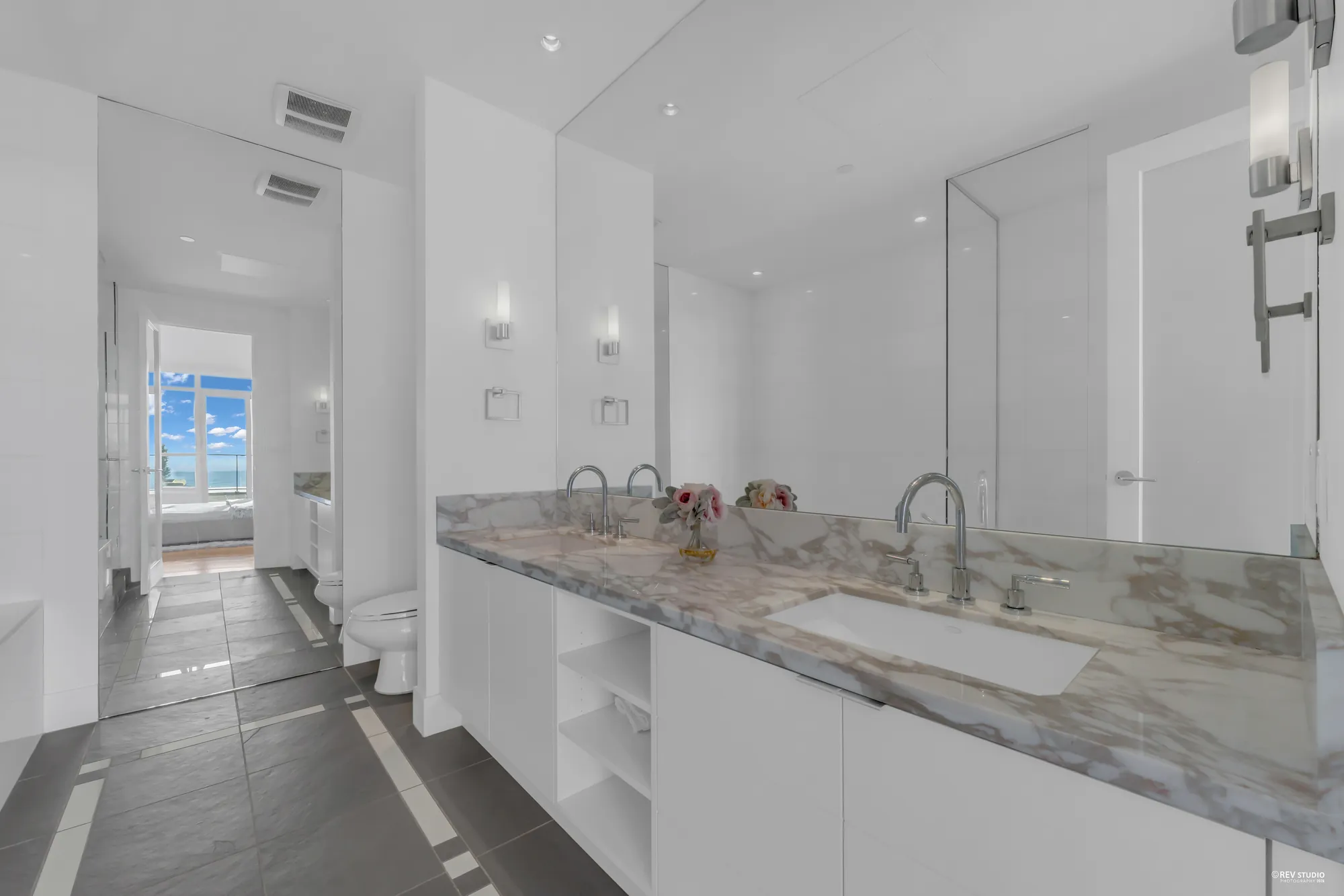 The primary bathroom: a long white vanity with two sinks and a veined stone counter, a glass shower and the toilet beyond, a grey tile floor