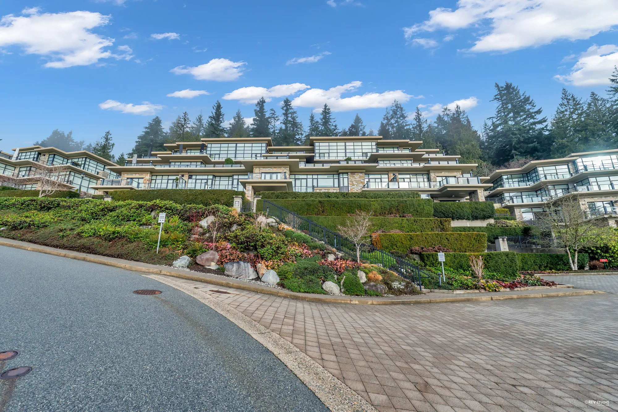 Whitby Estates from Twin Creek Place by day: terraced buildings of stone, concrete and glass stepping down a hillside of clipped hedges and rock gardens, evergreens behind, a paved turning circle in front