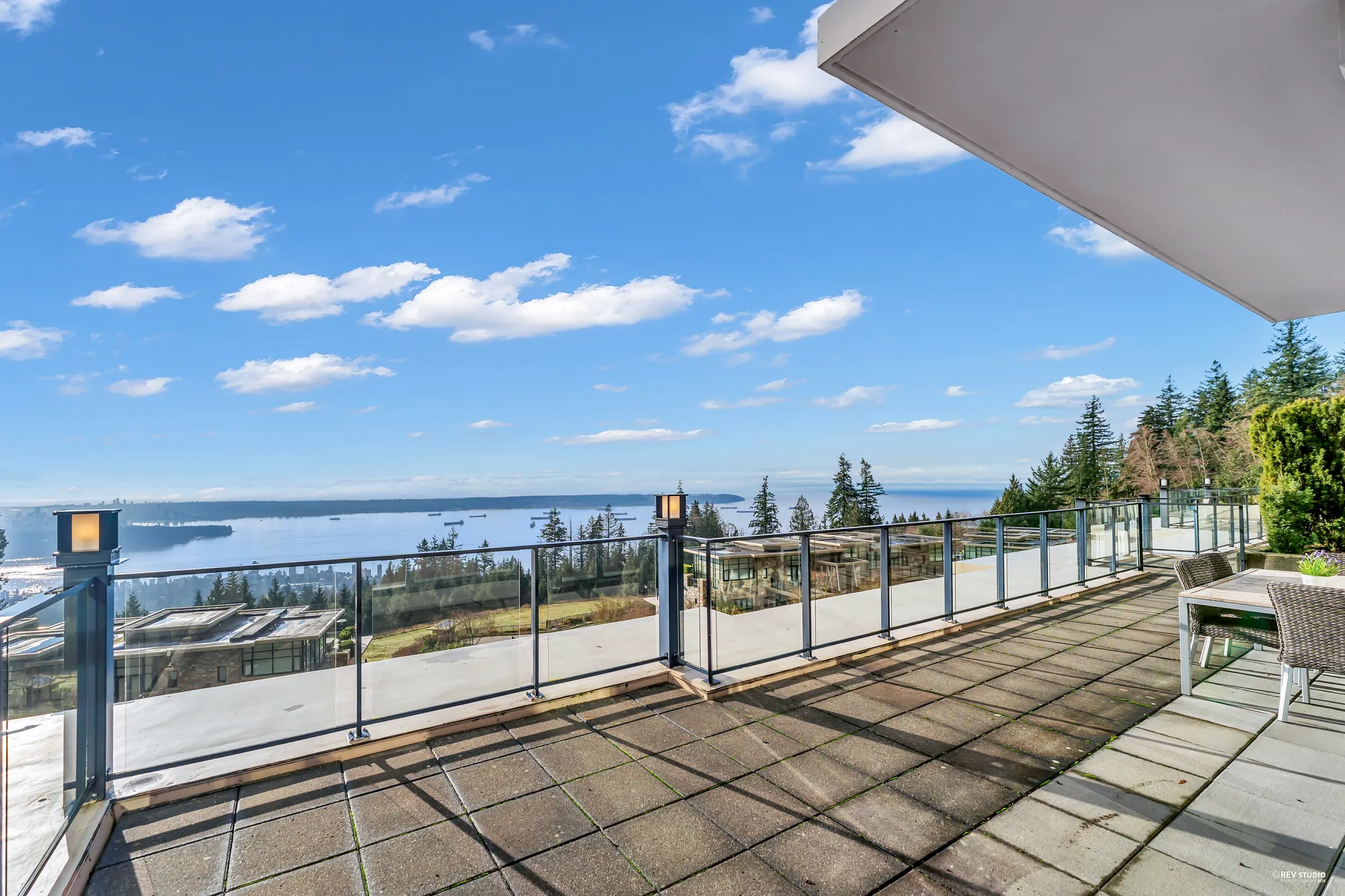 The terrace under the deep roof overhang: paving, a glass railing, and the water and the far shore under a blue sky