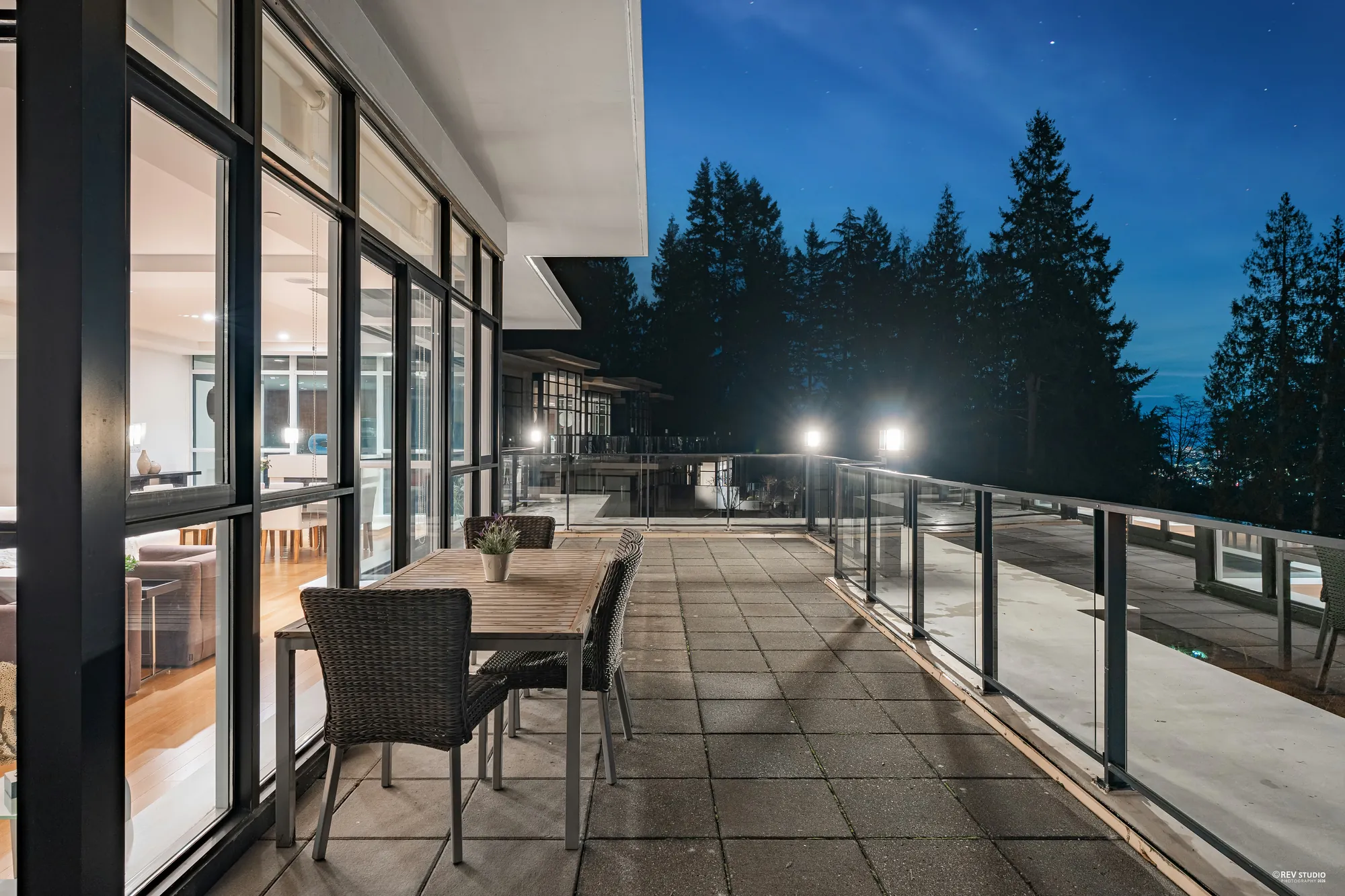 The terrace along the glass wall of the home at dusk, an outdoor dining table and chairs, lamps on the railing