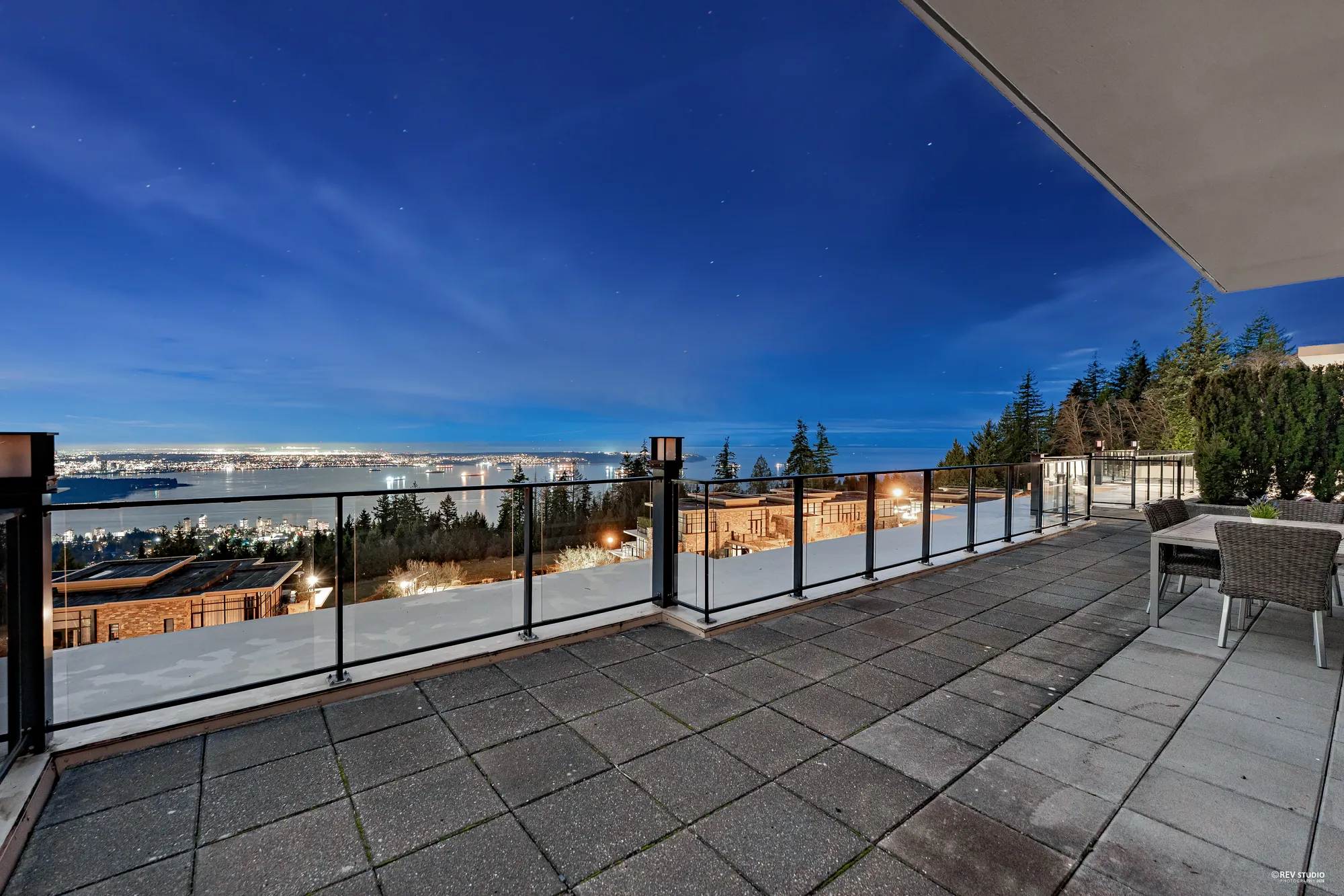 The terrace at dusk: the roof overhang and the glass railing in front, the water below and the lights of the city along the far shore under a deep blue sky